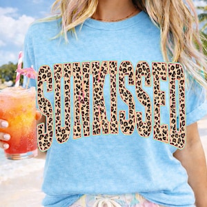 May include: A light blue short-sleeved t-shirt featuring the word SUNKISSED printed in a large, bold, block-letter font. The letters have a brown and black leopard print pattern with a thin, bright neon green outline around each character.
