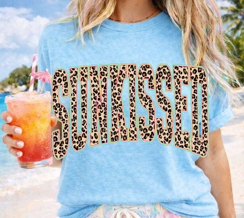 May include: A light blue short-sleeved t-shirt featuring the word SUNKISSED printed in a large, bold, block-letter font. The letters have a brown and black leopard print pattern with a thin, bright neon green outline around each character.