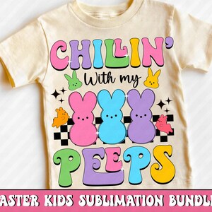 Puede incluir: Una camiseta crema para ni&ntilde;os con la frase "CHILLIN' With my PEEPS" en letras coloridas. El dise&ntilde;o incluye conejitos de dibujos animados y un patr&oacute;n de tablero de ajedrez, ideal para Pascua.