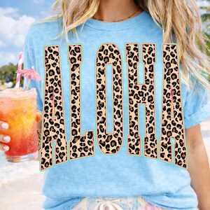 May include: Light blue t-shirt with the word "ALOHA" in large, leopard print letters. The letters have a teal outline. A tropical drink with a pink umbrella is held in the left hand.