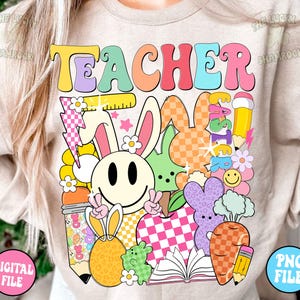 以下が含まれることがあります： 薄いベージュのTシャツに、カラフルなレトロな文字で「TEACHER」と書かれています。デザインには、笑顔のウサギ、チェック柄のリンゴ、本、その他カラフルな漫画の要素が描かれています。