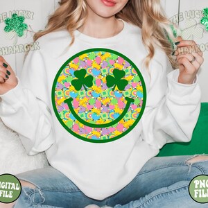 Peut inclure: Sweat-shirt blanc avec un motif de visage souriant vert. Le visage souriant est rempli de tr&egrave;fles, d'arcs-en-ciel et de pi&egrave;ces d'or color&eacute;s. Le texte "The Lucky Shamrock" est visible dans les coins sup&eacute;rieurs de l'image.