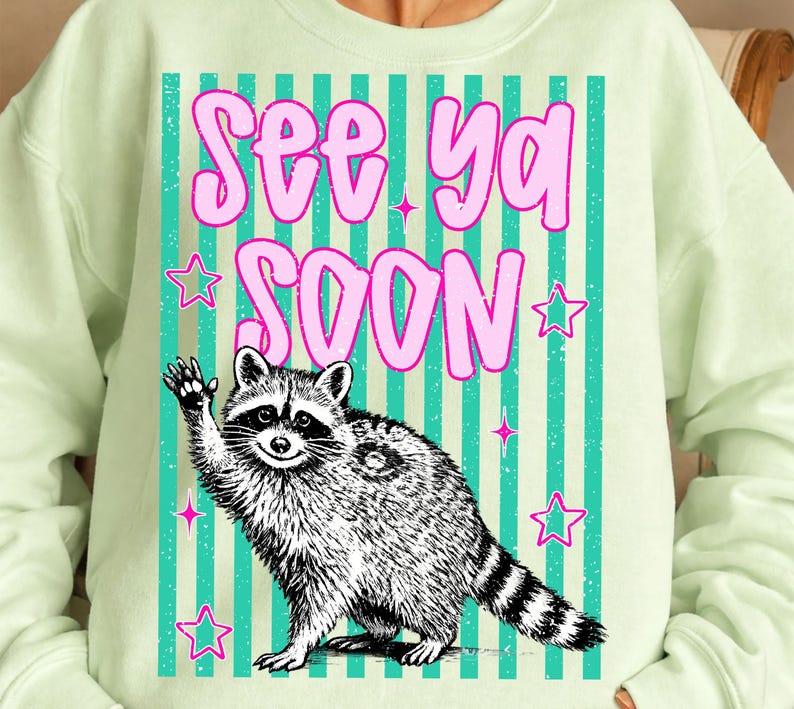 May include: A pale green sweatshirt with a raccoon illustration waving, accompanied by the text "See Ya Soon" in pink bubble letters. The design incorporates teal stripes and pink stars, offering a fun and quirky look.