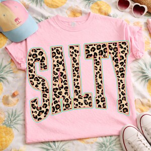 May include: A light pink t-shirt with the word "SALTY" in large, bold letters. The letters are filled with a leopard print design and outlined in a teal color. A baseball cap and sunglasses are also visible.