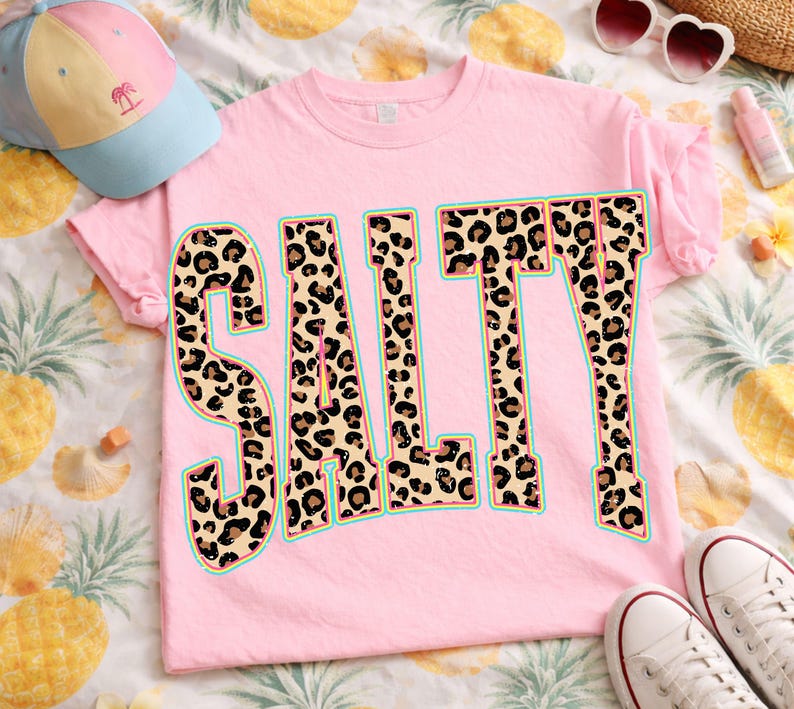May include: A light pink t-shirt with the word "SALTY" in large, bold letters. The letters are filled with a leopard print design and outlined in a teal color. A baseball cap and sunglasses are also visible.