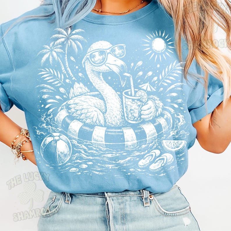 May include: Light blue t-shirt featuring a white graphic of a flamingo wearing sunglasses and floating in a pool with a drink. The design includes palm trees, a beach ball, and flip-flops, evoking a summer theme.