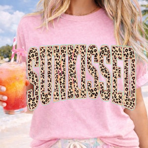 May include: A light pink short-sleeved t-shirt featuring the word SUNKISSED printed in large, bold capital letters. The letters have a tan, black, and brown leopard print pattern with a thin teal outline. The fabric has a textured, washed appearance.