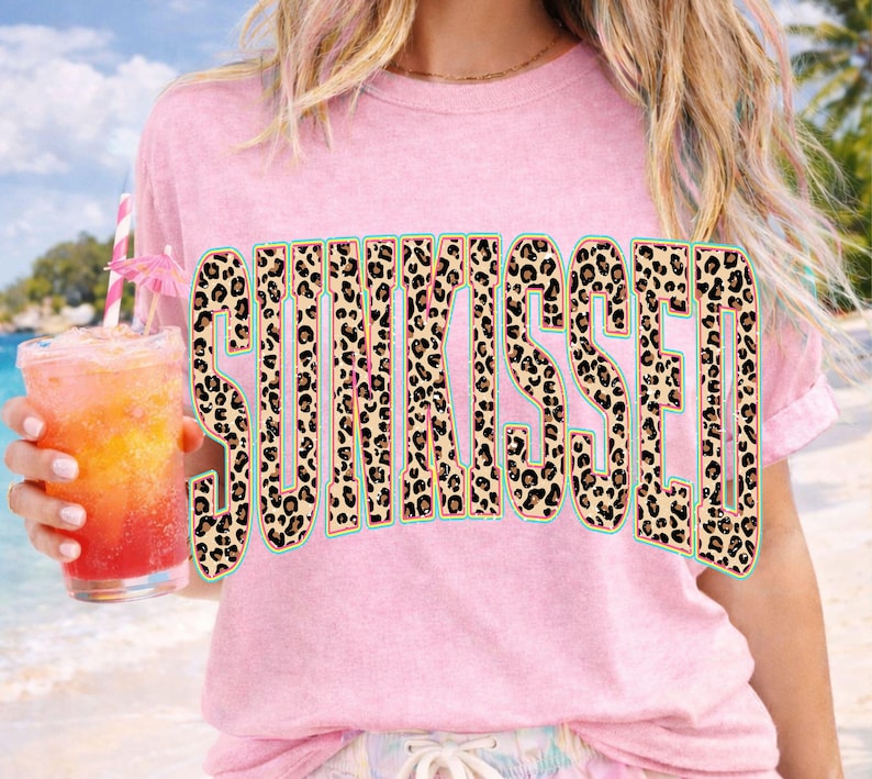 May include: A light pink short-sleeved t-shirt featuring the word SUNKISSED printed in large, bold capital letters. The letters have a tan, black, and brown leopard print pattern with a thin teal outline. The fabric has a textured, washed appearance.