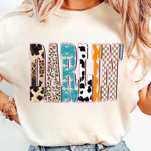May include: Cream-colored t-shirt with the word "DARLIN'" in a colorful, distressed font. The letters feature various patterns, including leopard print, turquoise, cow print, orange, and a checkered design, outlined in pink.
