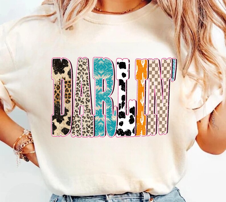 May include: Cream-colored t-shirt with the word "DARLIN'" in a colorful, distressed font. The letters feature various patterns, including leopard print, turquoise, cow print, orange, and a checkered design, outlined in pink.