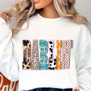 May include: White sweatshirt with the word "DARLIN" in a colorful, patterned design. The letters feature various textures, including leopard print, turquoise, and a checkered pattern. The sweatshirt is a casual, comfortable style.