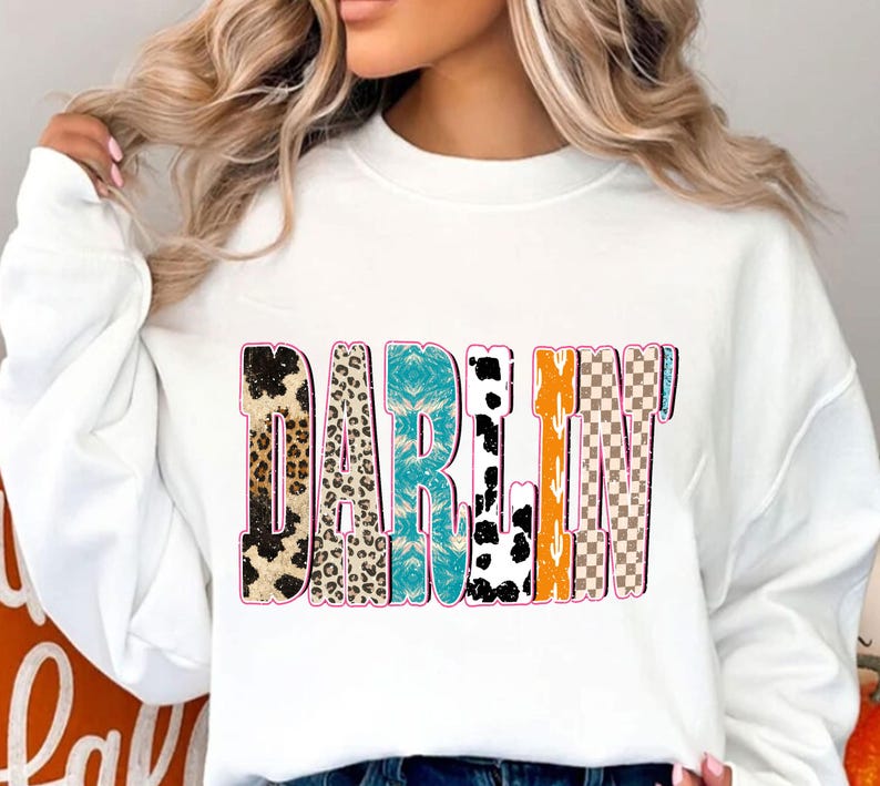 May include: White sweatshirt with the word "DARLIN" in a colorful, patterned design. The letters feature various textures, including leopard print, turquoise, and a checkered pattern. The sweatshirt is a casual, comfortable style.