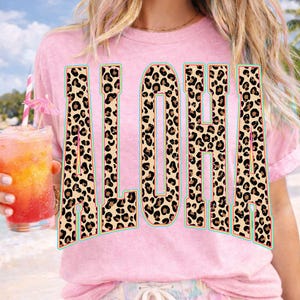 May include: Pink t-shirt with the word "ALOHA" in large, bold letters. The letters have a leopard print pattern with a teal outline. A tropical drink with a pink umbrella is held in the left hand.