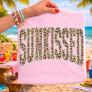 May include: A light pink t-shirt featuring the word SUNKISSED printed in a large, bold, block-letter font. The letters have a tan and black leopard print pattern with a thin teal outline, centered on the front of the garment.