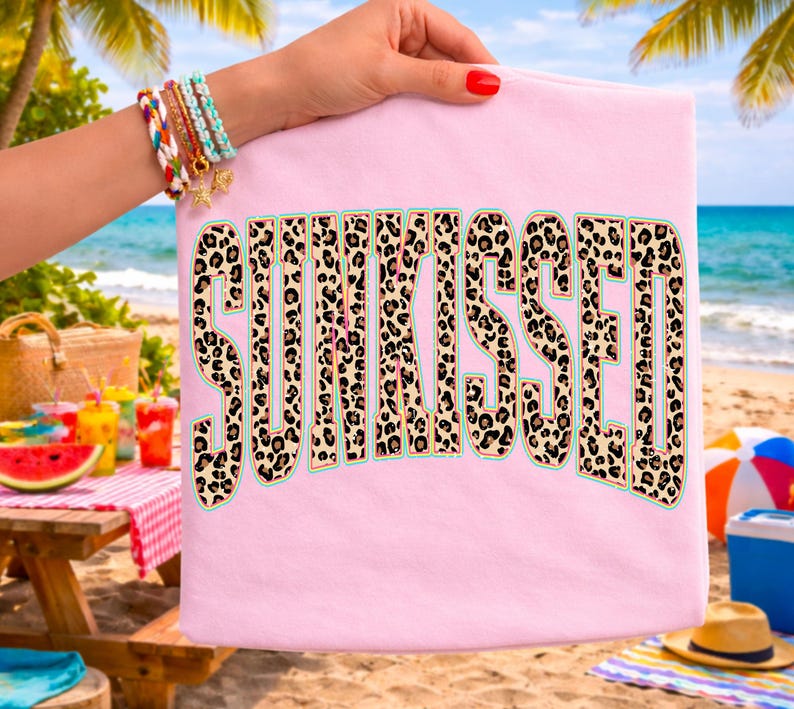 May include: A light pink t-shirt featuring the word SUNKISSED printed in a large, bold, block-letter font. The letters have a tan and black leopard print pattern with a thin teal outline, centered on the front of the garment.
