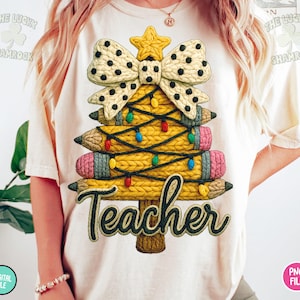 May include: Cream-colored t-shirt featuring a Christmas tree design made of pencils. The tree is adorned with a bow, star, and colorful lights. The word "Teacher" is written in a decorative font at the bottom of the tree.