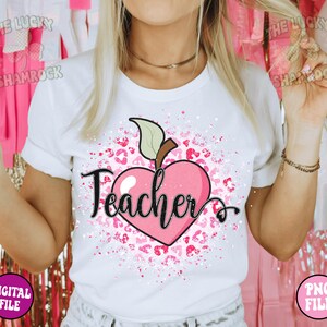 May include: White t-shirt with a pink heart-shaped apple and the word "Teacher" in black script. The apple is surrounded by pink leopard print and white splatters.