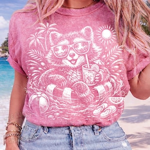 May include: Pink t-shirt featuring a white graphic of a raccoon wearing sunglasses and floating in a pool with a drink. The design includes palm trees, a beach ball, and the sun, evoking a summer vacation theme.