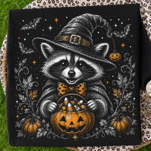 May include: A black t-shirt featuring a graphic illustration of a raccoon wearing a witch hat and a polka-dot bow tie. The raccoon holds a jack-o'-lantern bucket filled with candy corn. The design includes bats, stars, and pumpkins.