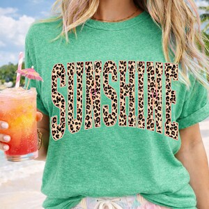 May include: A teal green t-shirt with the word "SUNSHINE" in large, leopard print letters. The shirt is worn by a person holding a tropical drink with a pink umbrella and a straw.