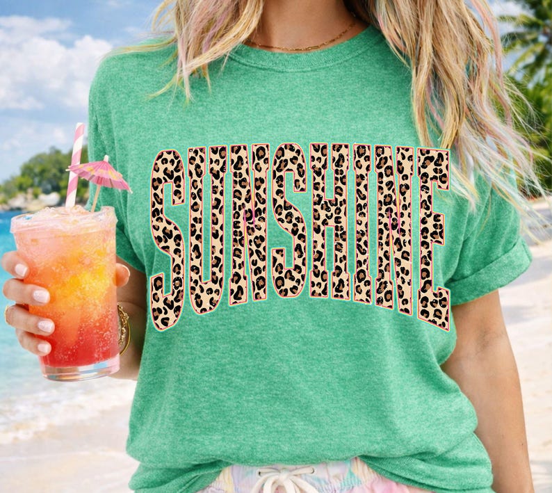 May include: A teal green t-shirt with the word "SUNSHINE" in large, leopard print letters. The shirt is worn by a person holding a tropical drink with a pink umbrella and a straw.