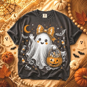 May include: A charcoal gray t-shirt featuring a graphic of a white ghost wearing a large orange polka-dot bow. The ghost is waving and holding an orange jack-o'-lantern bucket filled with candy. The design includes bats, stars, and a crescent moon.