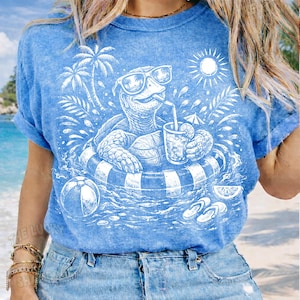 May include: Light blue t-shirt featuring a white graphic of a turtle wearing sunglasses and floating in a striped inner tube, holding a drink. The design includes palm trees, a sun, a beach ball, and flip-flops.