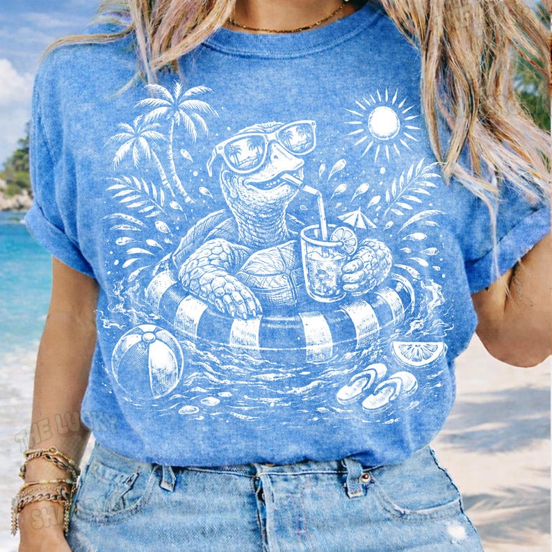 May include: Light blue t-shirt featuring a white graphic of a turtle wearing sunglasses and floating in a striped inner tube, holding a drink. The design includes palm trees, a sun, a beach ball, and flip-flops.