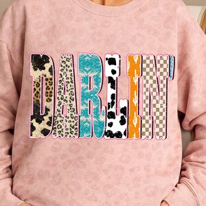 May include: A light pink sweatshirt with the word "DARLIN" in a colorful, patterned design. The letters feature various textures, including leopard print, turquoise, cow print, and a checkered pattern.