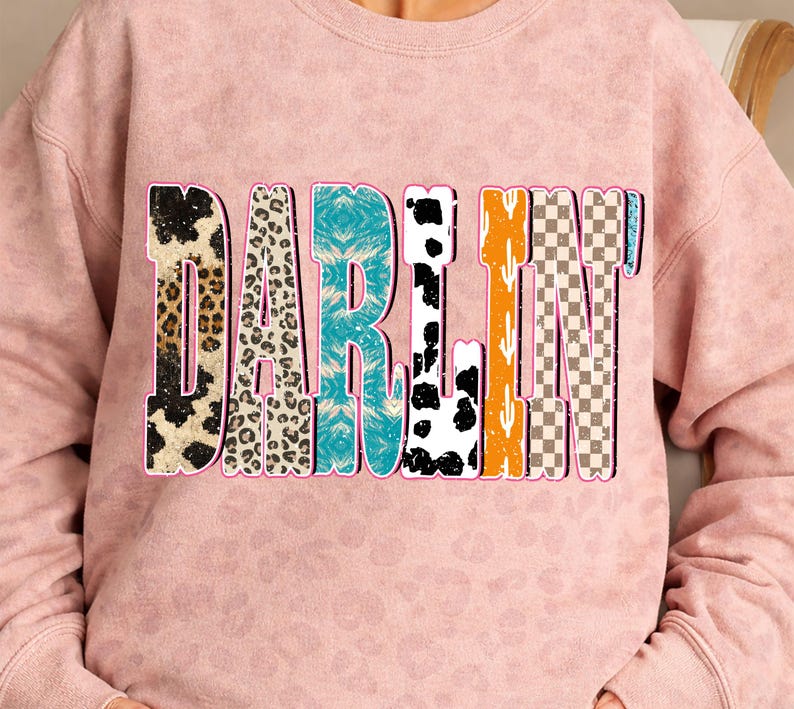 May include: A light pink sweatshirt with the word "DARLIN" in a colorful, patterned design. The letters feature various textures, including leopard print, turquoise, cow print, and a checkered pattern.