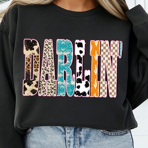 May include: Black crewneck sweatshirt with the word "DARLIN'" in a colorful, patterned design. The letters feature animal print, turquoise, cow print, orange cactus, and a checkered pattern, outlined in pink.