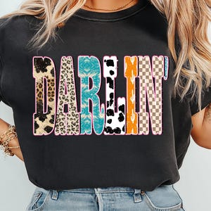 May include: Black t-shirt with the word "DARLIN" in a colorful, patterned design. The letters feature animal print, turquoise, black and white cow print, orange cactus, and a checkered pattern, all outlined in pink.