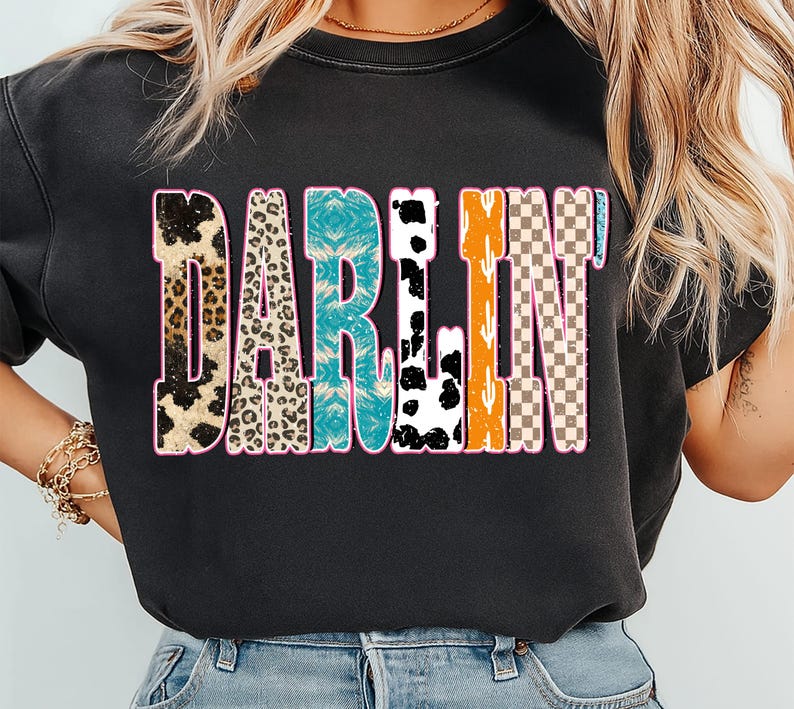 May include: Black t-shirt with the word "DARLIN" in a colorful, patterned design. The letters feature animal print, turquoise, black and white cow print, orange cactus, and a checkered pattern, all outlined in pink.