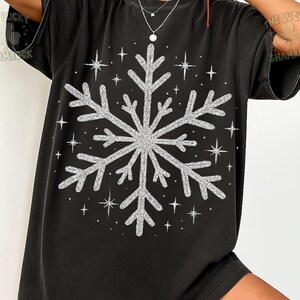 May include: Black t-shirt featuring a large, glittery silver snowflake design. Surrounding the snowflake are several smaller, sparkling star shapes. The shirt is a casual, oversized style, perfect for winter or holiday wear.
