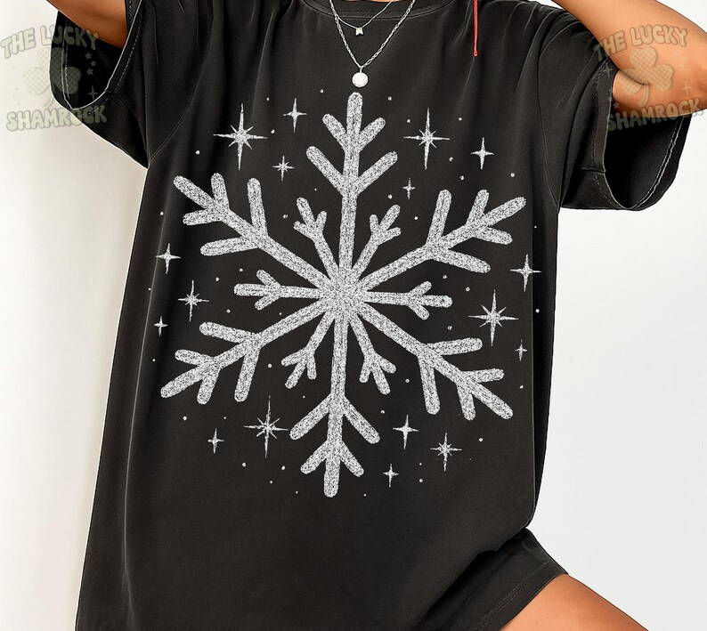 May include: Black t-shirt featuring a large, glittery silver snowflake design. Surrounding the snowflake are several smaller, sparkling star shapes. The shirt is a casual, oversized style, perfect for winter or holiday wear.