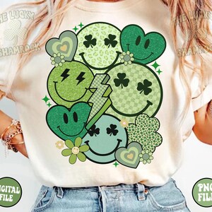 Peut inclure: Un t-shirt blanc avec un motif vert, blanc et noir de diff&eacute;rents visages souriants, c&oelig;urs et tr&egrave;fles. Le motif est de style r&eacute;tro avec des accents de paillettes.