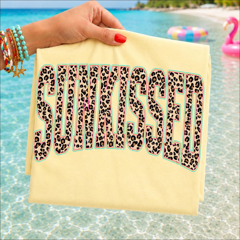 May include: A pale yellow t-shirt featuring the word SUNKISSED printed in large block letters. The letters have a brown and black leopard print pattern with a thin teal outline. The fabric is smooth and the design is centered on the front.