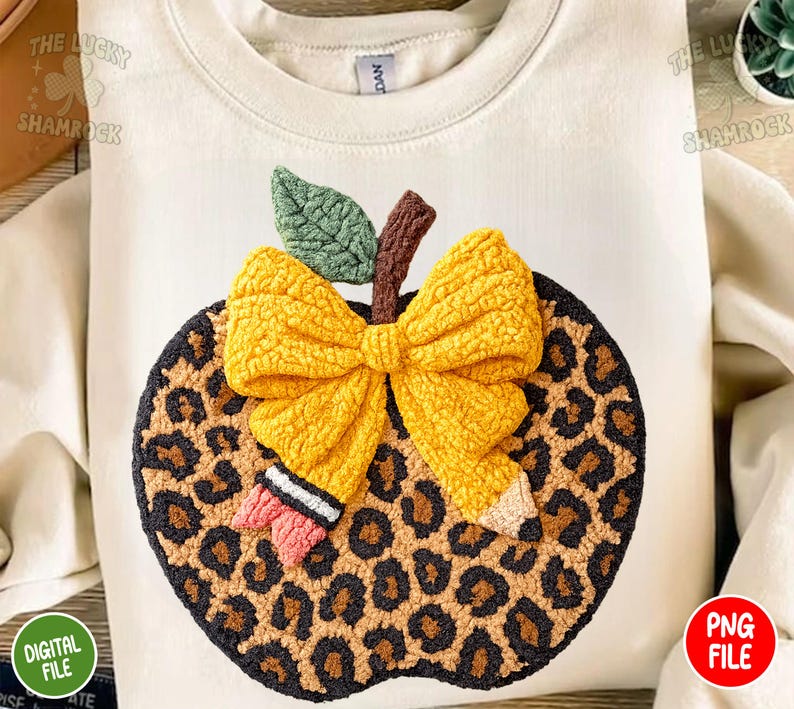 May include: Cream-colored sweatshirt featuring a large apple design. The apple has a leopard print pattern, a yellow bow, a green leaf, and a brown stem. A pencil is also part of the design. The words "DIGITAL FILE" and "PNG FILE" are visible.