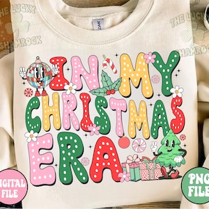 K&ouml;nnte beinhalten: Ein cremefarbener Pullover mit einem bunten Grafikdesign, auf dem "In My Christmas Era" steht. Die Grafik zeigt einen Weihnachtsbaum, Kugeln, Zuckerstangen und Geschenke.