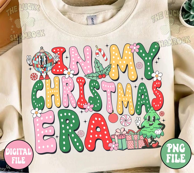 K&ouml;nnte beinhalten: Ein cremefarbener Pullover mit einem bunten Grafikdesign, auf dem "In My Christmas Era" steht. Die Grafik zeigt einen Weihnachtsbaum, Kugeln, Zuckerstangen und Geschenke.