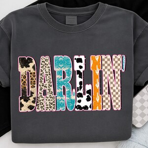 May include: Dark gray t-shirt with the word "DARLIN" in a colorful, patterned design. The letters feature animal prints, turquoise tie-dye, black and white cow spots, orange cacti, and a brown and white checkered pattern.