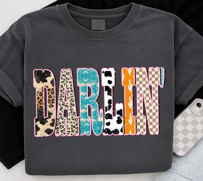 May include: Dark gray t-shirt with the word "DARLIN" in a colorful, patterned design. The letters feature animal prints, turquoise tie-dye, black and white cow spots, orange cacti, and a brown and white checkered pattern.