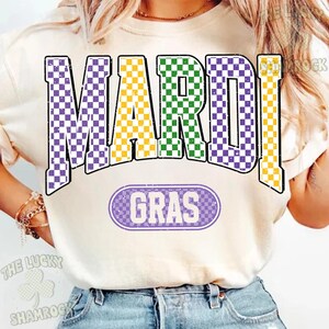 May include: A cream-coloured t-shirt with the words "MARDI GRAS" in a purple, yellow, and green chequered pattern. Below, in a purple oval, is the word "GRAS". The shirt is a casual style.