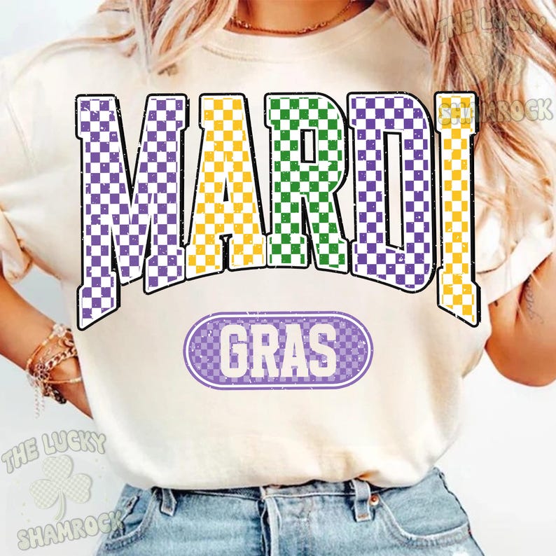 May include: A cream-coloured t-shirt with the words "MARDI GRAS" in a purple, yellow, and green chequered pattern. Below, in a purple oval, is the word "GRAS". The shirt is a casual style.
