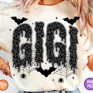 May include: Cream-colored t-shirt featuring the word "GIGI" in a dripping, leopard-print design. The shirt includes bat and spiderweb graphics, with bats and spiders. The design has a Halloween theme.