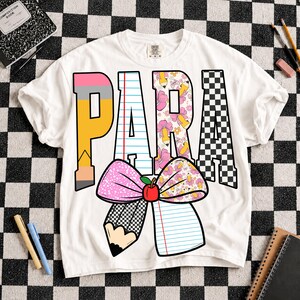 K&ouml;nnte beinhalten: Ein wei&szlig;es T-Shirt mit dem Wort PARA in gro&szlig;en Blockbuchstaben. Das P ist ein gelber Bleistift, das A ist liniertes Papier, das R hat ein Blumen-Bleistift-Muster und das A ist ein schwarz-wei&szlig;es Schachbrettmuster. Darunter ist eine gro&szlig;e Schleife mit einem roten Apfel.