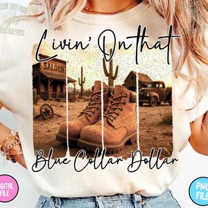 Puede incluir: Camiseta color crema con un gr&aacute;fico de tem&aacute;tica western. El gr&aacute;fico incluye botas de trabajo marrones, un saloon, una camioneta y el texto "Livin' On that Blue Collar Dollar". La camiseta tiene mangas cortas y cuello redondo.