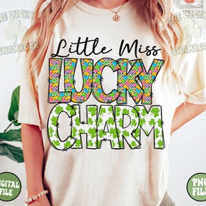 Peut inclure: Un t-shirt blanc avec le texte "Little Miss Lucky Charm" en lettres noires soulign&eacute;es. Le mot "Lucky" est d&eacute;cor&eacute; d'un motif de tr&egrave;fles &agrave; quatre feuilles color&eacute;s. Le mot "Charm" est d&eacute;cor&eacute; de motifs de tr&egrave;fles &agrave; quatre feuilles verts.