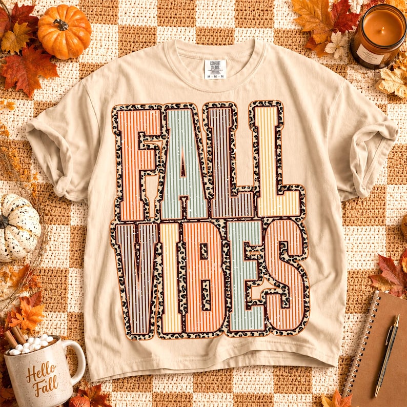 May include: A beige short-sleeve t-shirt featuring the text FALL VIBES in large block letters. The letters have a vertical striped pattern in muted orange, blue, and yellow, outlined with a brown and black leopard print border.