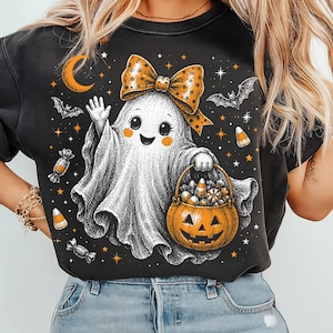 May include: A black crewneck sweatshirt featuring a graphic of a white ghost with a large orange polka-dot bow, waving one hand and holding a jack-o'-lantern bucket filled with candy. The design includes bats, stars, a crescent moon, and candy.